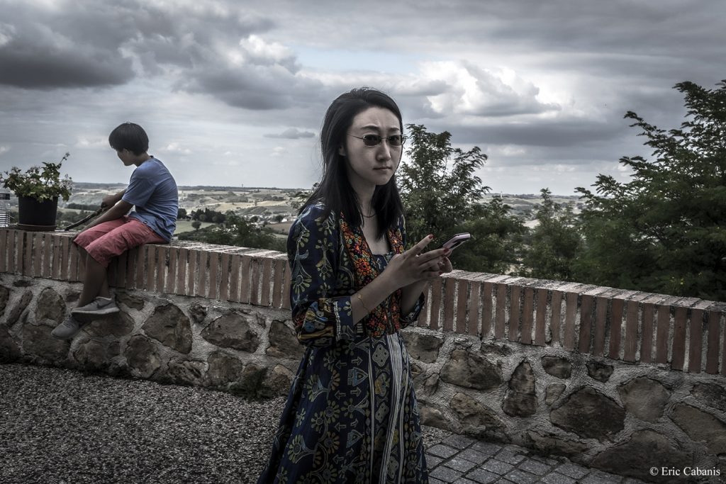 Une jeune femme marche près des remparts du Carla-Bayle , une cité d'artistes en Ariège, son téléphone à la main Photojournalisme Streetphotography Eric Cabanis