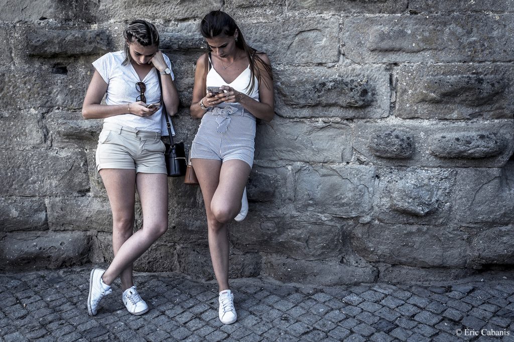 Deux jeunes femmes regardent leurs téléphones portables adossées aux remparts de la cité médiévale de Carcassonne en été Photojournalisme Streetphotography Eric Cabanis