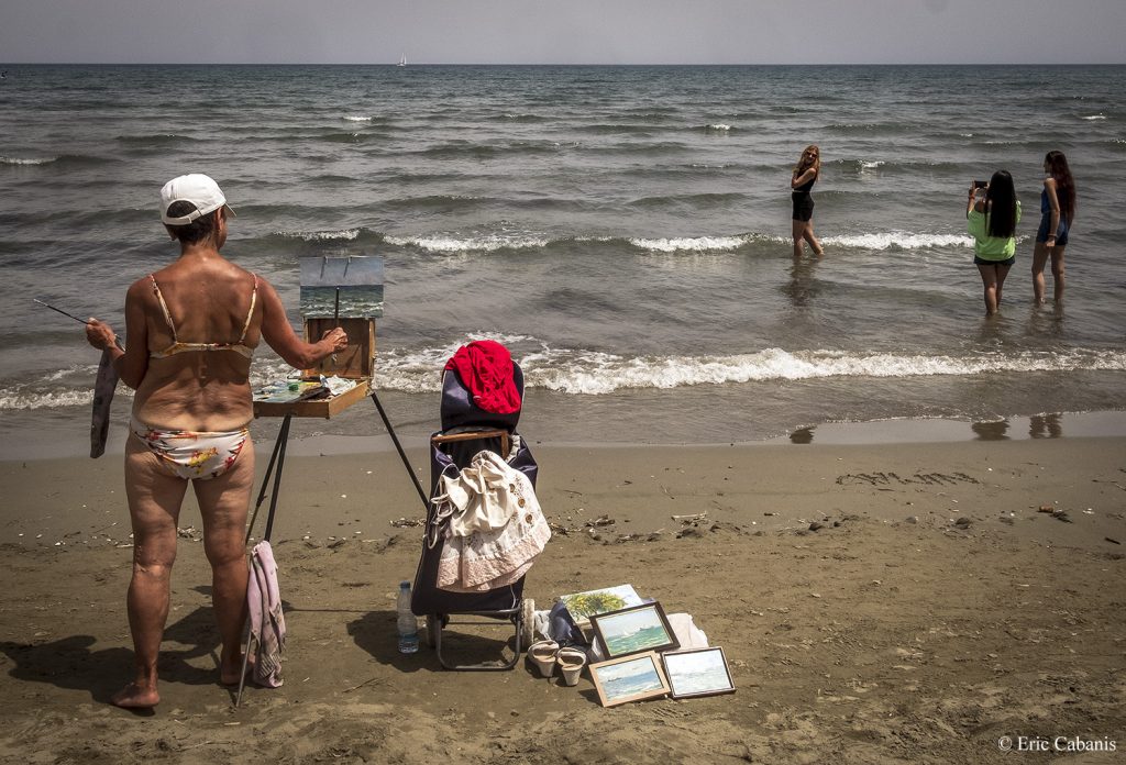 Sur la plage de Larnaca à Chypre en mai 2018 une femme peint la mer pendant que des jeunes filles se prennent en photo dans l'eau Photojournalism Streetphotography Eric Cabanis