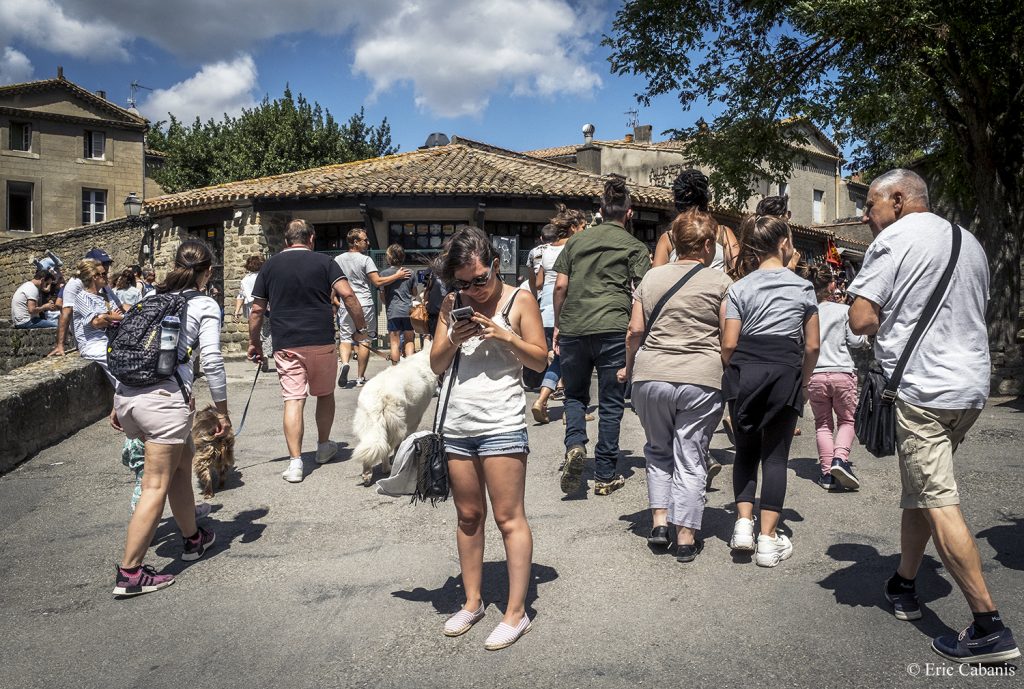 Une jeune femme consulte son téléphone portable au milieu des nombreux touristes de la cité médiévale de Carcassonne en août 2019 Photojournalisme Streetphotography Eric Cabanis